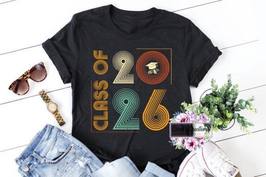 Black t-shirt with 'Class of 2026' design on a white wooden floor with accessories.