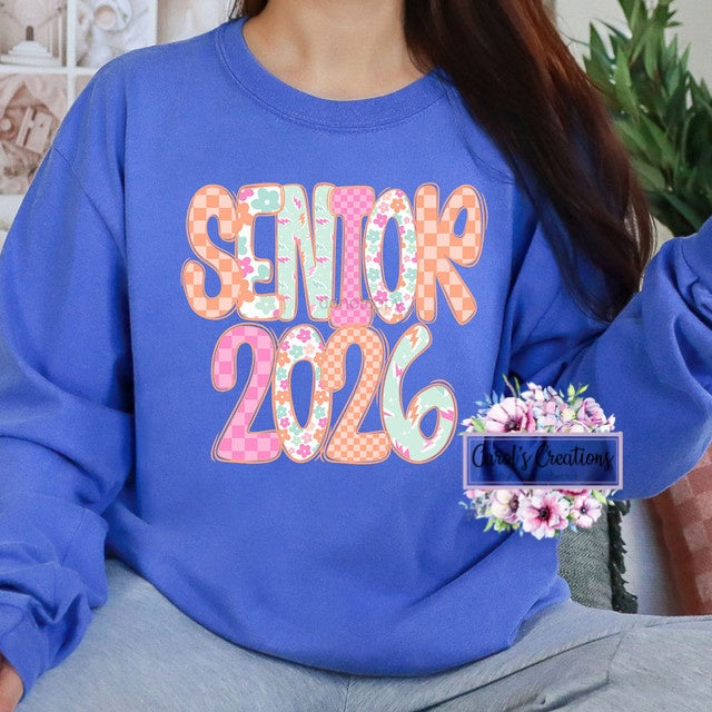 Person wearing a blue sweatshirt with 'Senior 2026' text and floral design, sitting indoors.